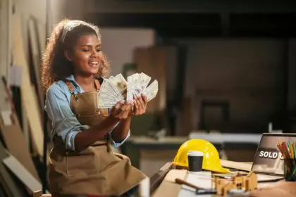 Attractive black woman carpenter holding banknote from selling wood furniture in small business.