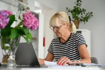 Old woman wearing glasses working on laptop online, sitting at desk on living room