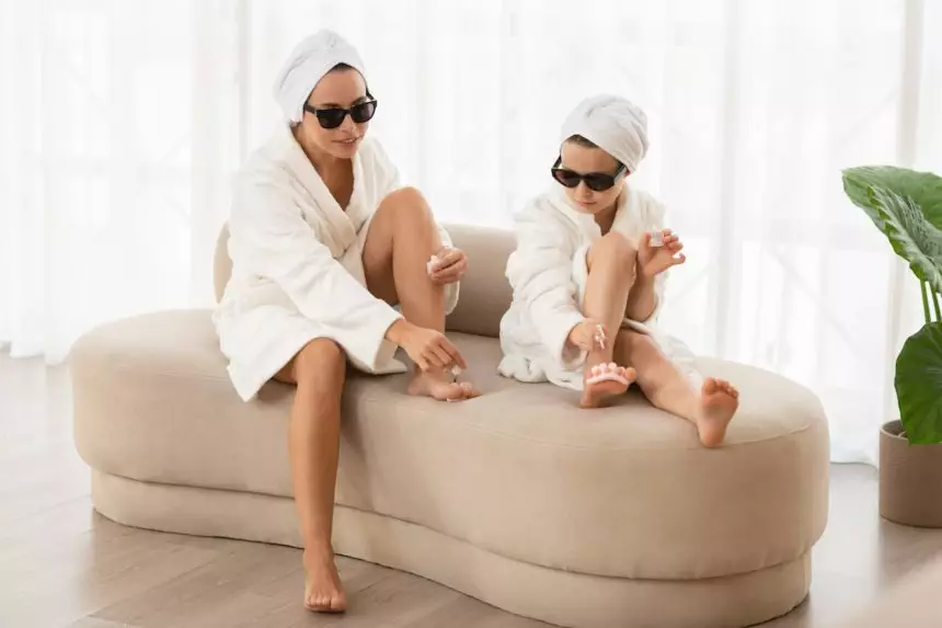 Mother and little daughter in bathrobes and sunglasses making pedicure at home,