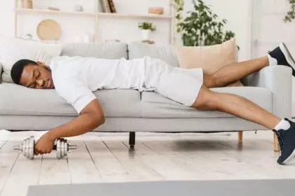 Exhausted Black Man Holding Dumbbell Sleeping On Sofa At Home
